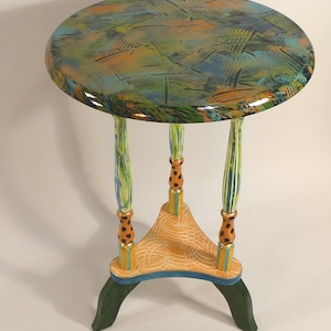 May include: A small, round side table with a colorful, abstract design. The tabletop features a mix of green, blue, and orange hues. The table has three legs, each with a unique painted pattern, and a triangular base. The table is approximately 24 inches tall.