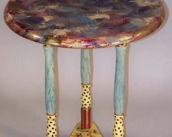 May include: A small, round, wooden table with a colorful, abstract painted top. The table has three legs, each painted with a blue, yellow, and red striped pattern and black polka dots.