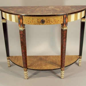 May include: A half-round, wooden console table with a single drawer and a lower shelf. The table has a black base with gold accents and painted legs with a black, white, and red striped pattern. The top of the table is a dark brown with gold accents.