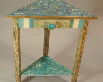 May include: A triangular wooden table with a painted blue and green top and a lower shelf. The table has a single drawer with a blue knob and a white and green striped border.