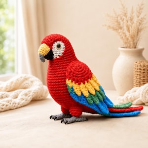 Puede incluir: Una figura de loro tejida a ganchillo con un cuerpo rojo vibrante, un parche facial blanco y un pico amarillo y negro. Las alas muestran plumas en capas amarillas, verdes y azules, mientras que las plumas de la cola son verdes, rojas y azules. Pies grises.