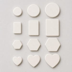 Puede incluir: Una colección de doce formas de cerámica blanca dispuestas en una cuadrícula. El conjunto incluye tres círculos, tres rectángulos, tres hexágonos y tres corazones, cada uno presentado en tres tamaños diferentes de pequeño a grande con acabado mate.