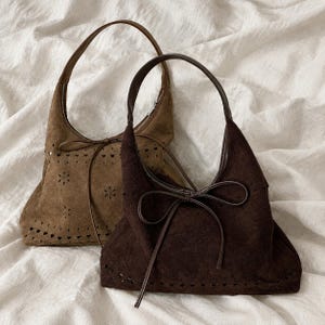 May include: Two hobo-style shoulder bags in dark brown and tan suede. Each bag features a single handle, a front tie closure, and a decorative laser-cut pattern of small flowers and hearts along the lower section and the upper border.