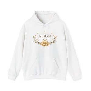 May include: A white hooded sweatshirt featuring a central graphic print. The design includes the word ALIGN in brown capital letters, positioned above a golden crescent moon and a lotus flower, surrounded by delicate leafy branches and stars.