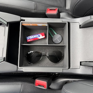 Peut inclure: Un plateau organisateur de console centrale de voiture en plastique gris foncé avec trois compartiments. Le plateau contient un paquet de chewing-gum Stimorol, un petit appareil électronique rond noir avec une sangle verte et des lunettes aviateur noires.