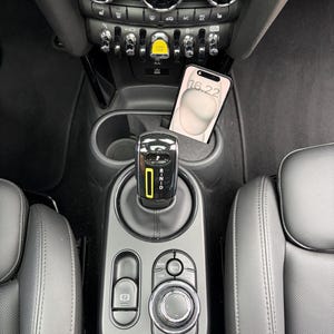 Peut inclure: La console centrale intérieure d'un véhicule comprenant un levier de vitesses noir avec un accent vertical jaune, une molette de commande circulaire, un bouton de frein de stationnement et un smartphone placé dans un porte-gobelet affichant 16:22.