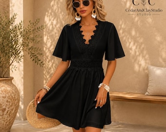 May include: A black mini dress featuring a deep V-neckline with intricate lace trim, short flutter sleeves, and a fitted waist with a matching lace band. The dress has a flared skirt and is accented by a logo in the top right corner reading CedarAndClayStudio.