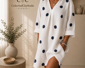 May include: A white V-neck dress featuring a pattern of dark blue polka dots, three-quarter length sleeves, and side pockets. The garment is displayed with the text CedarAndClayStudio in the upper left corner, indicating the brand name.