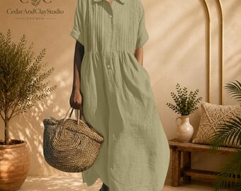 May include: A sage green midi dress with a collared neckline, short sleeves, and a button-down front. The dress features vertical pleats on the bodice and a loose, flowing skirt. A woven straw tote bag is held at the side of the garment.