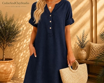 May include: A navy blue knee-length dress with a V-neckline, a partial button-down front featuring three light-coloured buttons, short sleeves with folded cuffs, and side pockets. The dress is displayed with a tan woven semi-circular handbag.