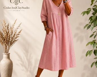 May include: A dusty pink midi dress featuring a V-neckline, three-quarter length sleeves, and vertical lace trim detailing along the front. The dress includes side pockets and a loose, flowing silhouette. The text CedarAndClayStudio is visible.