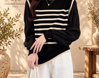 May include: A long-sleeved jumper featuring a black and cream horizontal striped knit bodice with a round neckline. The sleeves are made of a solid black, textured fabric with gathered cuffs. The hem and neckline are finished with cream trim.