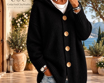 May include: A black hooded cardigan featuring a chunky knit texture, long sleeves, and four large, round, light-colored wooden buttons down the front. The garment is worn over a white shirt and paired with light blue distressed denim jeans.