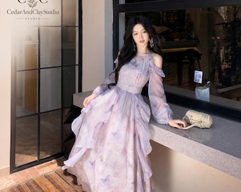 May include: A long-sleeved, cold-shoulder dress in a light lavender hue, featuring a layered, ruffled skirt and a delicate butterfly print. The garment has a fitted waist, long sheer sleeves with cuffs, and a high neckline. CedarAndClayStudio logo.