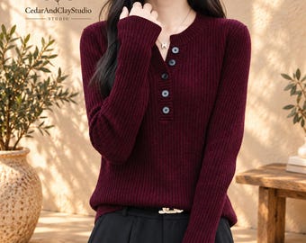 May include: A long-sleeved, ribbed knit jumper in a deep burgundy colour. The jumper features a round neckline with a partial button-down placket containing five dark buttons. The garment is paired with black trousers featuring a gold-toned belt buckle.