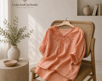 May include: A peach-coloured blouse featuring a V-neckline with intricate tonal embroidery around the collar and chest. The garment has short sleeves with buttoned cuffs and a relaxed, flowing silhouette, displayed on a wooden hanger.