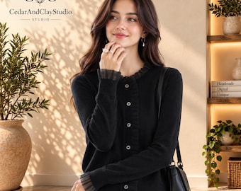 May include: A black long-sleeved cardigan featuring a round neckline, a front button closure, and delicate sheer mesh ruffles at the cuffs and collar. The garment is paired with a black shoulder bag and light-coloured trousers.