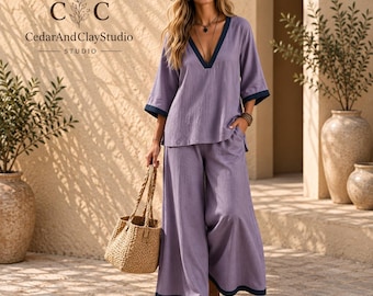 May include: A lavender two-piece set consisting of a V-neck top with three-quarter sleeves and wide-leg trousers, both featuring dark navy blue trim. The outfit is paired with a woven straw tote bag and a wide-brimmed straw hat.