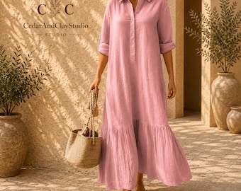 May include: A long-sleeved, floor-length dress in a soft pink hue, featuring a collared neckline, a button-down front placket, and a tiered skirt design. The garment is made of a textured fabric and is paired with a woven straw tote bag.