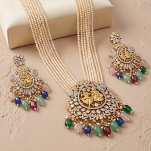 May include: A multi-strand pearl necklace featuring a large circular gold-tone pendant with a central figure motif, surrounded by clear crystals and dangling multi-coloured gemstone beads, accompanied by matching drop earrings with similar designs.