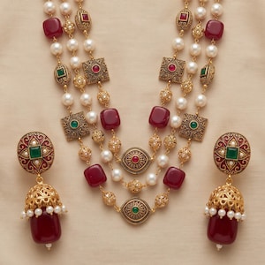 Può includere: Una collana a tre fili con perle bianche, perline in filigrana color oro e pietre quadrate rosse e verdi. Il set include orecchini abbinati in stile jhumka con pietre centrali verdi e perline rosse pendenti con dettagli in perla.