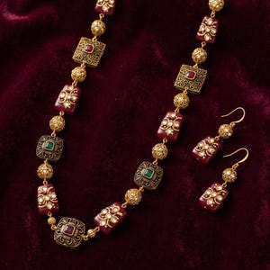 May include: A gold-toned necklace and matching drop earrings set featuring alternating maroon and green rectangular stones, intricate gold filigree square accents, and small gold textured spherical beads strung along a gold-toned chain.