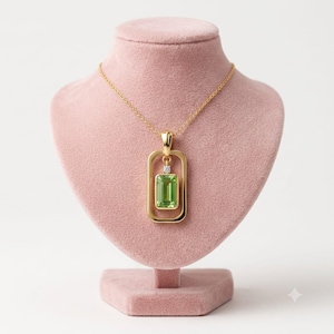 Puede incluir: Un collar con colgante de oro que presenta una piedra preciosa rectangular de color verde claro con corte esmeralda suspendida dentro de un marco rectangular de oro. Una pequeña piedra redonda transparente se sitúa sobre la gema verde.
