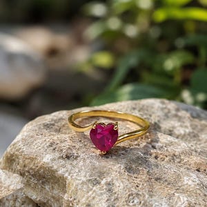 Puede incluir: Un anillo de oro con una piedra preciosa de color rosa vibrante en forma de corazón, montada en un engaste de garras. La banda es lisa y pulida, con un diseño sencillo y fino que se curva ligeramente hacia la piedra central en forma de corazón.