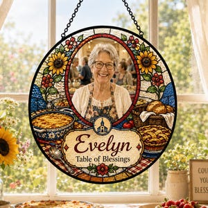 May include: A circular stained glass suncatcher featuring a central portrait of a woman, surrounded by sunflowers, berries, and food illustrations. The text reads Evelyn Table of Blessings with a small church icon, suspended by a black chain.
