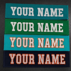 May include: Four rectangular embroidered patches arranged vertically. The top two patches are teal and green with white text reading YOUR NAME. The bottom two are teal and navy with orange text reading YOUR NAME in a block varsity font style.