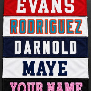 May include: Five rectangular fabric patches with embroidered text. The top is red with white text EVANS, followed by white with orange-outlined blue text RODRIGUEZ, navy with white text DARNOLD, white with navy text MAYE, and black with pink text YOUR NAME.