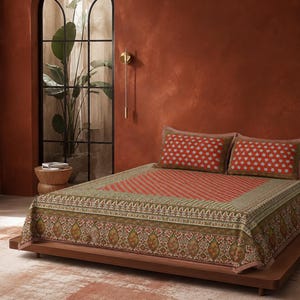 Könnte beinhalten: Eine Tagesdecke mit einem zentralen orangefarbenen Feld mit weißem Muster, umgeben von einer breiten olivgrünen und braunen Bordüre mit komplexen floralen und geometrischen Motiven. Zwei passende rechteckige Kissen liegen am Kopfende.