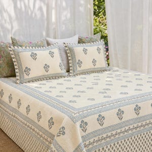 May include: A cream-colored bedspread and two matching rectangular pillow shams featuring a repeating blue floral block print pattern. The bedding set includes a decorative border with intricate blue geometric and floral motifs on the edges.