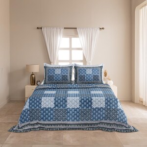 Op de afbeelding: Een blauw-witte patchwork quiltset met een herhalend raster van gevarieerde geometrische en bloemmotieven. De set bevat een sprei met een geplooide rand en twee bijpassende rechthoekige kussenslopen, die allemaal een consistent indigo-kleurig blokprintontwerp over het gehele textieloppervlak vertonen.