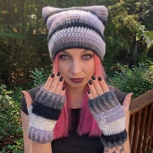 Puede incluir: Un gorro de ganchillo con orejas de gato y guantes sin dedos a juego, con un patrón de rayas horizontales en tonos morado claro, gris, blanco y negro. El gorro tiene dos orejas puntiagudas y los guantes presentan un diseño de puño acanalado.
