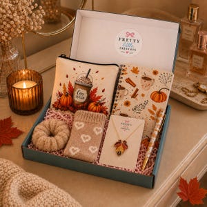 May include: A gift box labelled Pretty Little Presents containing a zippered pouch with a coffee graphic, a spiral notebook with autumn leaves and pumpkins, a beige heart-patterned sock, a gold-tone leaf pendant necklace, and a patterned pen.