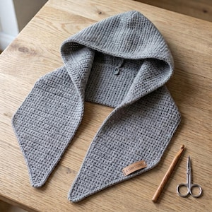 May include: A light gray crocheted hood with two long, pointed scarf-like ends. The hood features a textured stitch pattern and a small brown rectangular leather tag with the text A YARN STUDIO. A wooden crochet hook and metal scissors are nearby.