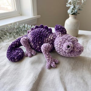 May include: A plush chameleon toy crafted from soft, velvety yarn in shades of deep purple and light lavender. The stuffed animal features a textured, crocheted body with a curled tail, a row of small dorsal spikes, and two black plastic eyes.