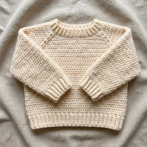 May include: A cream-colored long-sleeve sweater featuring a textured crochet knit pattern. The garment has a round neckline, raglan sleeves, and ribbed cuffs at the wrists and hemline. The construction displays a consistent stitch throughout.