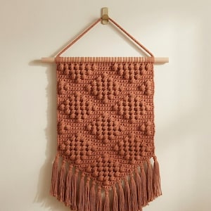 May include: A terracotta-colored crochet wall hanging featuring a textured bobble stitch pattern in a diamond arrangement. The piece is suspended from a light-colored wooden dowel with a matching yarn cord and finished with a fringe at the bottom.