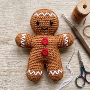 Könnte beinhalten: Eine braune, gehäkelte Lebkuchenfigur mit zwei roten Knöpfen auf der Brust, weißer Zickzack-Stickerei an Kopf, Armen und Beinen sowie schwarzen Kunststoffaugen. Die Figur hat ein aufgesticktes schwarzes Lächeln und eine runde Kopfform.