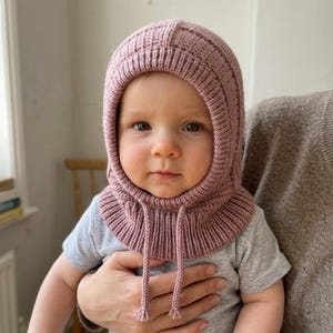 May include: A dusty rose knitted balaclava hood with a ribbed texture and a wide, circular collar. The headpiece features a snug fit around the face and two thin, braided ties hanging from the front, finished with small, knotted tassels at the ends.