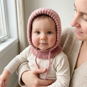 May include: A dusty rose knitted bonnet featuring a textured ribbed pattern around the face and a ribbed collar base. The headwear includes two thin matching knitted ties hanging down from the front, crafted in a soft yarn material.