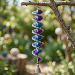 May include: A spiral-shaped hanging decoration crafted from crocheted yarn in alternating shades of purple, blue, and light blue. The structure features a beige yarn border and terminates in a small wooden bead and a light beige yarn tassel.