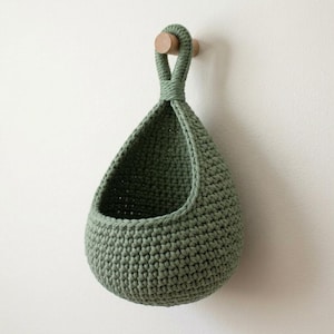 May include: A sage green crocheted hanging basket with a teardrop shape and a single integrated loop handle. The basket is constructed from thick yarn in a consistent stitch pattern and is suspended from a small, round wooden wall hook.
