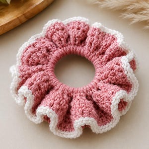 May include: A circular hair scrunchie made of dusty rose pink yarn with a textured, ruffled appearance. The outer edge of the scrunchie is finished with a delicate, scalloped trim in a contrasting off-white yarn, creating a layered effect.