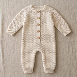 Peut inclure: Une barboteuse à manches longues de couleur crème en fil crocheté. Le vêtement présente une encolure ronde, des poignets et des chevilles côtelés, ainsi qu'une patte de boutonnage verticale sur le devant avec cinq petits boutons en bois clair.