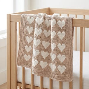 Op de afbeelding: Een rechthoekige gehaakte deken met een herhalend patroon van witte hartjes op een beige achtergrond. De deken heeft een effen beige rand met een gestructureerd steekpatroon en hangt over de houten spijlen van een lichtgekleurd ledikant.