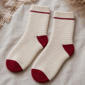May include: A pair of cream-colored crochet socks featuring a ribbed cuff with a thin horizontal red stripe, a solid red heel, and a solid red toe. The socks are crafted with a consistent stitch pattern throughout the main body of the fabric.