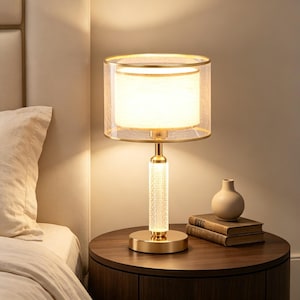 May include: A table lamp featuring a cylindrical gold base with a textured glass column and a dual-layer drum shade. The inner shade is opaque white, while the outer layer is a sheer, fine mesh material. The lamp emits a warm, soft glow.